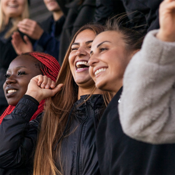 Trois jeunes femmes souriantes se tiennent côte à côte, exprimant de la joie et de l'enthousiasme. Elles portent des vestes noires, et l'une d'elles a des tresses rouges. En arrière-plan, d'autres personnes applaudissent, créant une ambiance festive.