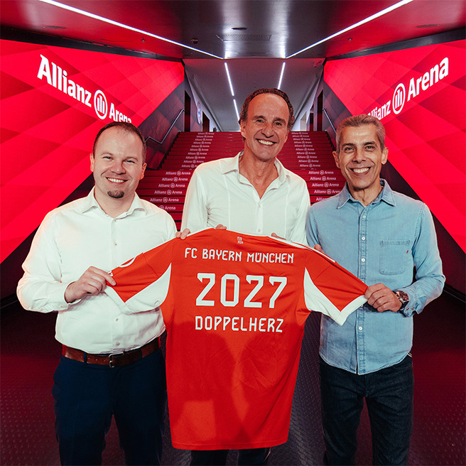 Trois hommes se tiennent au milieu de l'Allianz Arena, tenant un maillot de football rouge du FC Bayern Münchens avec "2027" et "DOPPELHERTZ" inscrits au dos. L'environnement est éclairé par des lumières rouges et des affiches du stade.