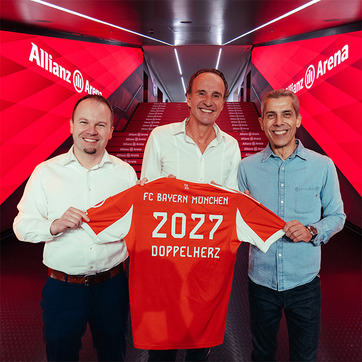 Trois hommes se tiennent au milieu de l'Allianz Arena, tenant un maillot de football rouge du FC Bayern Münchens avec "2027" et "DOPPELHERTZ" inscrits au dos. L'environnement est éclairé par des lumières rouges et des affiches du stade.