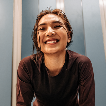 Une femme souriante avec les cheveux mouillés se tient devant un mur bleu. Elle porte un top sombre et semble heureuse, exprimant de l'énergie et de la confiance. L'image transmet une impression de joie et de dynamisme.