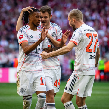 Des joueurs de football du Bayern Munich célèbrent un but. Un joueur au centre, vêtu d'un maillot blanc avec des détails rouges, lève les mains en signe de joie. Les autres joueurs l'encouragent, affichant des sourires et de l'enthousiasme dans un stade rempli de supporters.